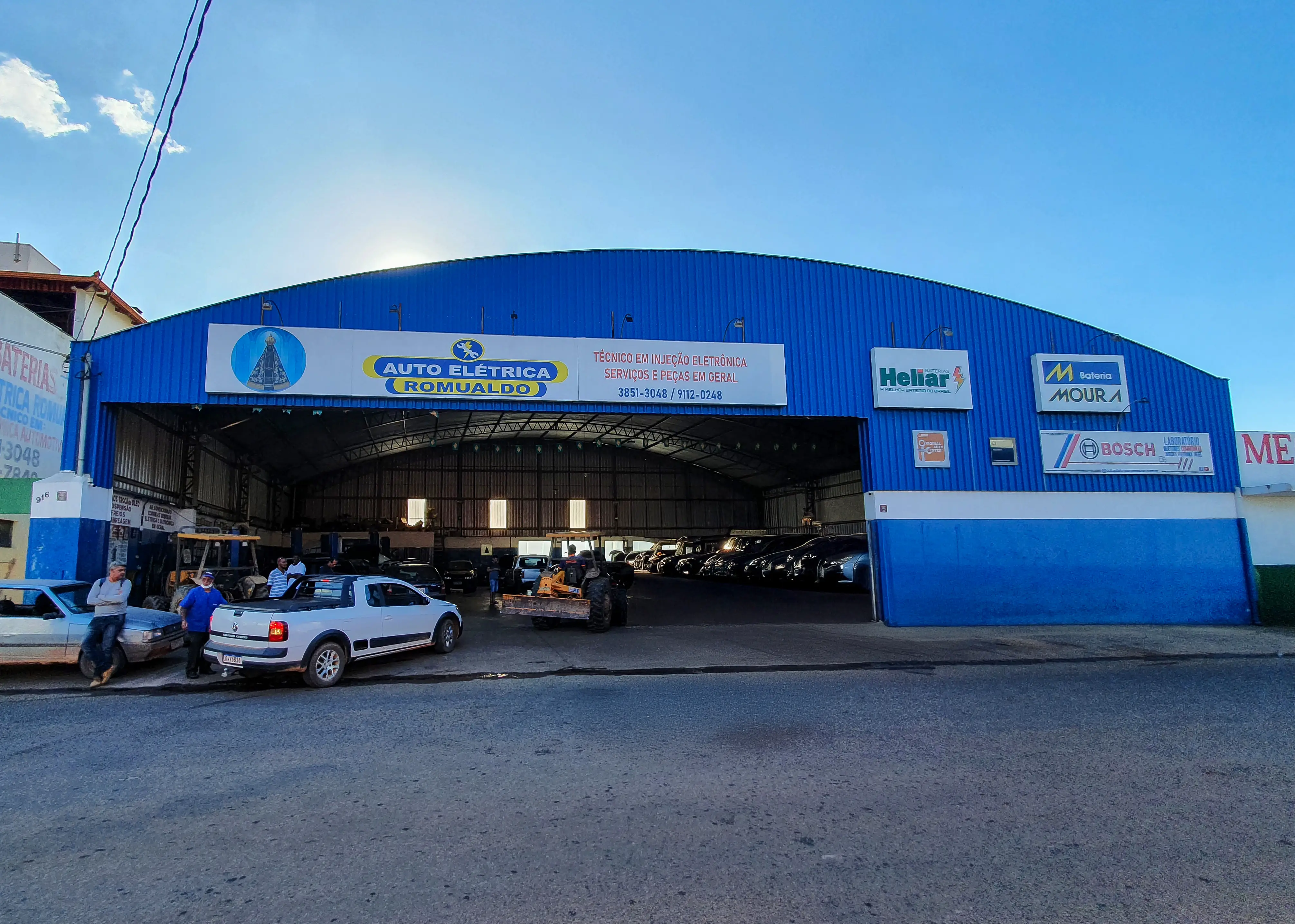 Fachada da Romualdo Autoelétrica & Autopeças em Boa Esperança/MG: galpão azul com o letreiro da oficina, veículos e equipe no pátio